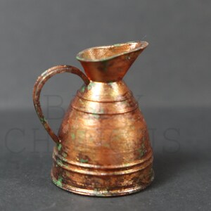 May include: A small copper pitcher with a green patina. The pitcher has a rounded body and a flared spout. It has a handle that is attached to the body with a decorative loop.