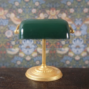 Puede incluir: Una lámpara de escritorio de vidrio verde con una base de metal dorado. La lámpara tiene una pantalla con forma de rectángulo y una parte superior redondeada.