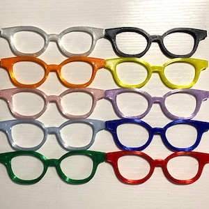 May include: A collection of ten miniature eyeglass frames arranged in two columns and five rows. The frames are solid colors including white, black, orange, yellow, light pink, purple, light blue, dark blue, green, and red, all featuring round lenses.