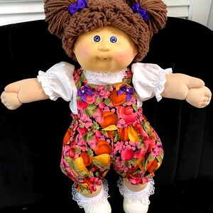 Puede incluir: Una mu&ntilde;eca con cabello de lana marr&oacute;n peinado en dos mo&ntilde;os con lazos morados, ojos azules y mejillas rosadas. Viste una camisa blanca de manga corta con encaje, un mameluco con estampado de frutas y lazos morados, calcetines con encaje y zapatos.