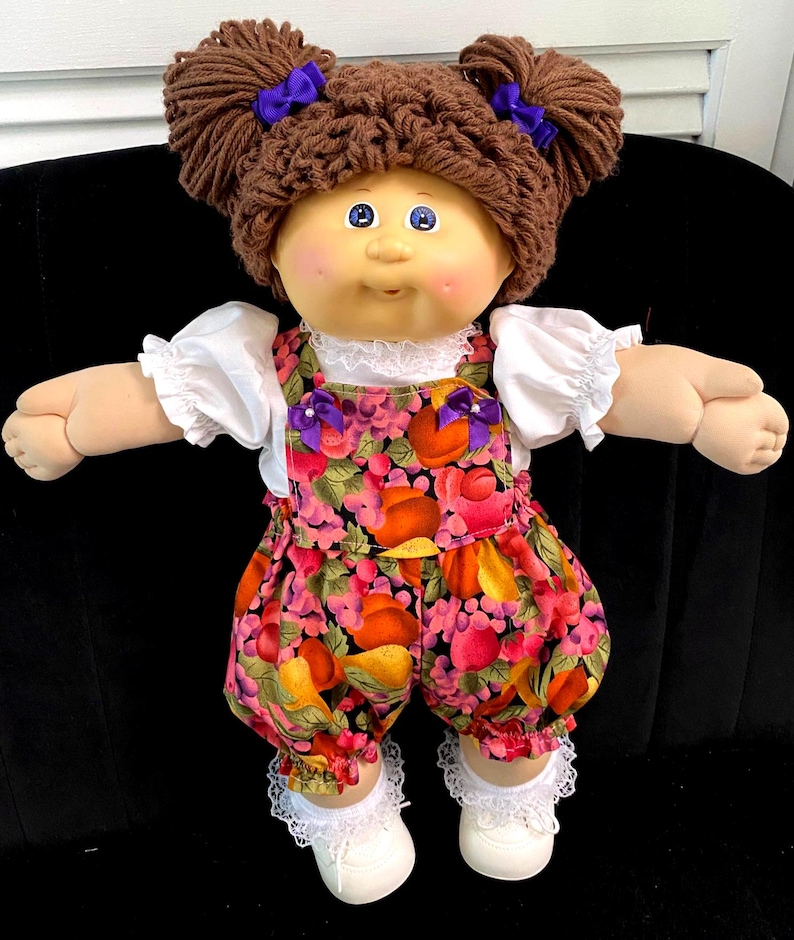 Puede incluir: Una mu&ntilde;eca con cabello de lana marr&oacute;n peinado en dos mo&ntilde;os con lazos morados, ojos azules y mejillas rosadas. Viste una camisa blanca de manga corta con encaje, un mameluco con estampado de frutas y lazos morados, calcetines con encaje y zapatos.