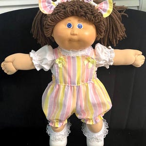Puede incluir: Una mu&ntilde;eca con cabello de lana marr&oacute;n peinado en coletas con lazos a cuadros, ojos azules y cara redonda. Viste una blusa blanca de manga corta con ribete de encaje y un pelele de rayas pastel con lazos amarillos en los hombros y zapatos.