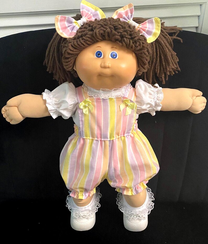 Puede incluir: Una mu&ntilde;eca con cabello de lana marr&oacute;n peinado en coletas con lazos a cuadros, ojos azules y cara redonda. Viste una blusa blanca de manga corta con ribete de encaje y un pelele de rayas pastel con lazos amarillos en los hombros y zapatos.