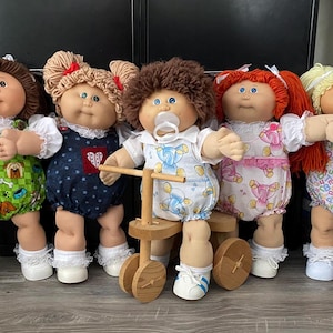 May include: Five Cabbage Patch Kids dolls with different hairstyles and outfits. One doll is riding a wooden tricycle. The dolls have various colored hair, including brown, red, and blonde. They wear rompers with patterns like dogs, hearts, and apples.