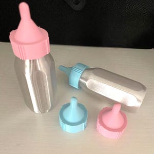 May include: Two silver-colored baby bottles with faceted sides, one standing upright with a pink nipple and cap, and one lying horizontally with a light blue nipple and cap. Two additional separate bottle caps with nipples in pink and light blue.