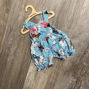Puede incluir: Un pelele azul claro con estampado de flores blancas y mariposas multicolores, con dos lazos de cinta rosa en los tirantes y aberturas el&aacute;sticas en las piernas, presentado en una percha de madera con el texto QuteZe.etsy.com impreso.