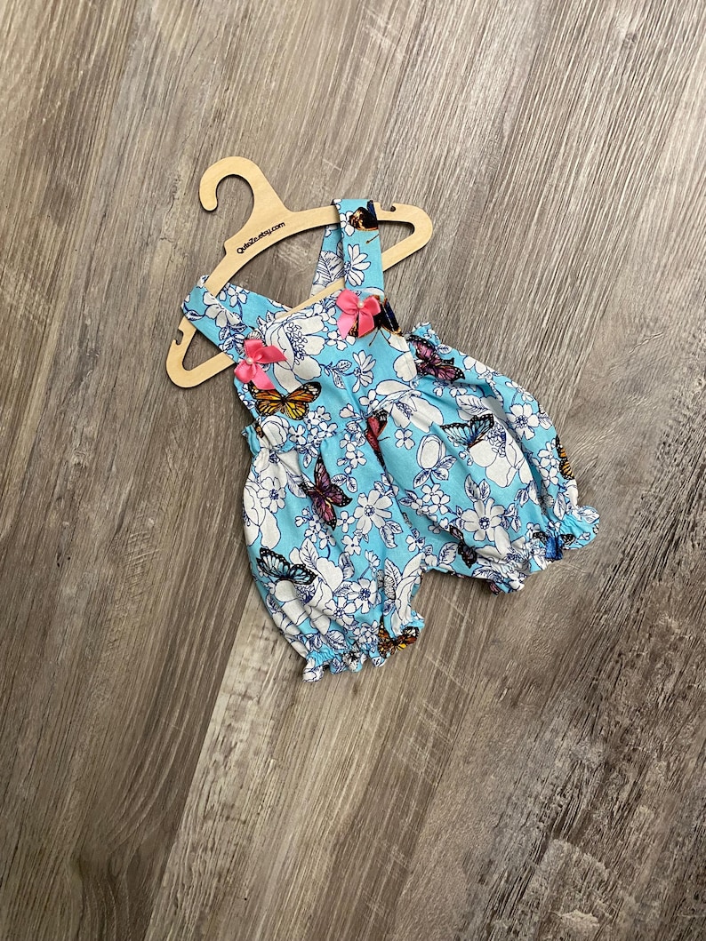 Puede incluir: Un pelele azul claro con estampado de flores blancas y mariposas multicolores, con dos lazos de cinta rosa en los tirantes y aberturas el&aacute;sticas en las piernas, presentado en una percha de madera con el texto QuteZe.etsy.com impreso.