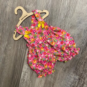 Puede incluir: Un pelele rosa con un vibrante estampado floral de flores amarillas, naranjas y blancas con tallos verdes. La prenda tiene aberturas de pierna con volantes, dos lazos de cinta amarilla en los tirantes y se muestra en una percha de madera.