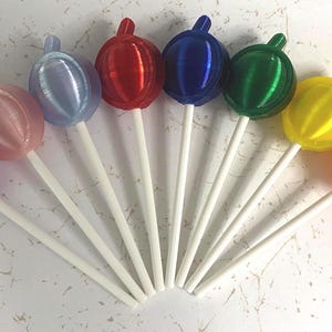 May include: A collection of nine spherical lollipops arranged in a fan shape, each featuring a smooth, glossy finish in a distinct color including purple, pink, light blue, red, dark blue, green, yellow, and orange, all attached to white sticks.
