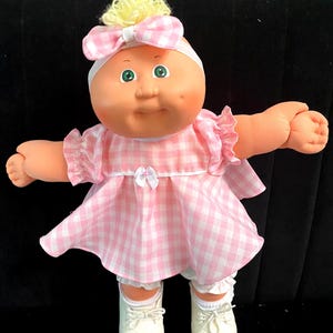 May include: A doll with a round face, green eyes, and blonde yarn hair styled in a top knot. The doll wears a pink and white gingham dress with a matching headband bow, white ruffled bloomers, and white lace-up shoes with a soft fabric body.
