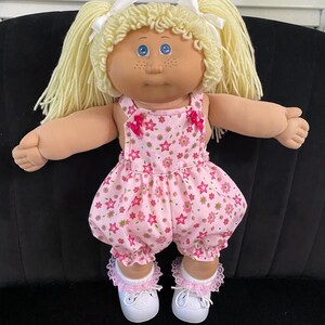 Puede incluir: Una mu&ntilde;eca de piel clara, ojos azules y pecas, con cabello de lana rubia peinado en dos coletas con cintas blancas. La mu&ntilde;eca viste un pelele rosa con estampado de flores y estrellas, calcetines blancos con encaje rosa y zapatos blancos.
