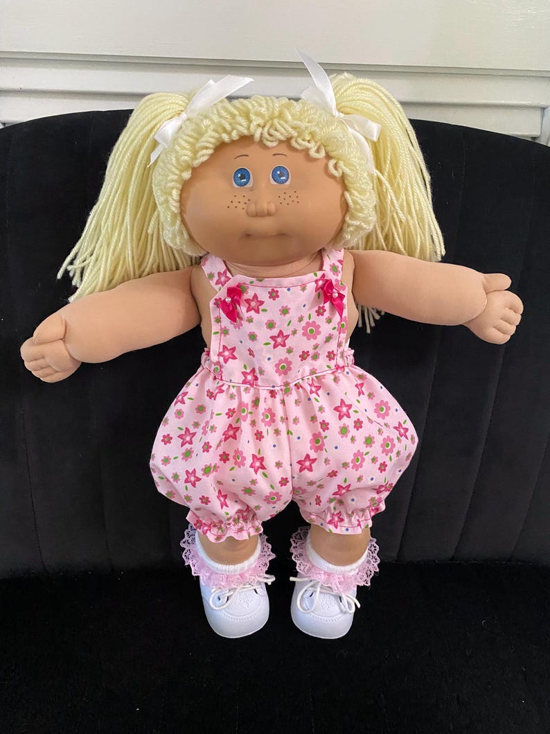 Puede incluir: Una mu&ntilde;eca de piel clara, ojos azules y pecas, con cabello de lana rubia peinado en dos coletas con cintas blancas. La mu&ntilde;eca viste un pelele rosa con estampado de flores y estrellas, calcetines blancos con encaje rosa y zapatos blancos.
