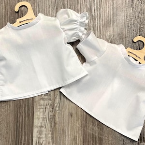 May include: Two white baby shirts on wooden hangers. One shirt has short sleeves with ruffled cuffs, and the other has short sleeves. The shirts are displayed on a wooden surface.