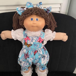 Puede incluir: Una mu&ntilde;eca con cabello de lana marr&oacute;n peinado en coletas con lazos azules de mariposas. La mu&ntilde;eca viste una blusa blanca de manga corta con encaje bajo un peto azul con estampado de mariposas y flores, decorado con dos lazos rosas.