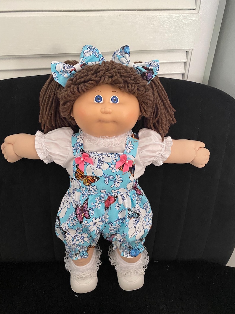 Puede incluir: Una mu&ntilde;eca con cabello de lana marr&oacute;n peinado en coletas con lazos azules de mariposas. La mu&ntilde;eca viste una blusa blanca de manga corta con encaje bajo un peto azul con estampado de mariposas y flores, decorado con dos lazos rosas.