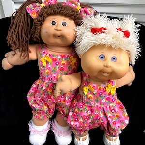 May include: Two dolls with round faces and soft bodies wearing matching pink floral overalls with yellow bows. The left doll has brown yarn hair with a matching bow, while the right doll has short white yarn hair with two small red bows.