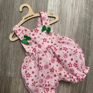 Puede incluir: Un pelele de color rosa claro con un estampado floral repetitivo en tonos rosa oscuro, verde claro y blanco. La prenda tiene tirantes, aberturas el&aacute;sticas en las piernas y dos peque&ntilde;os lazos de sat&eacute;n verde con cuentas blancas.