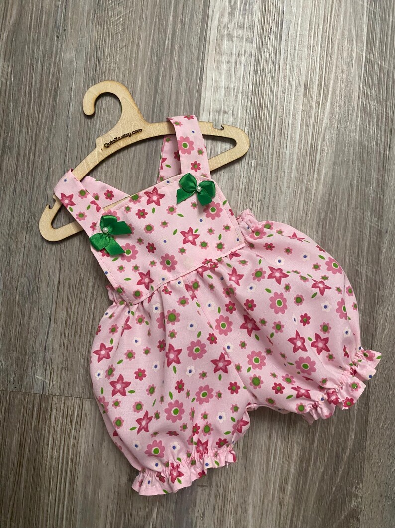 Puede incluir: Un pelele de color rosa claro con un estampado floral repetitivo en tonos rosa oscuro, verde claro y blanco. La prenda tiene tirantes, aberturas el&aacute;sticas en las piernas y dos peque&ntilde;os lazos de sat&eacute;n verde con cuentas blancas.