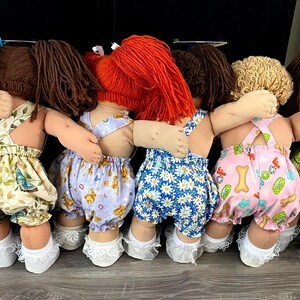 Puede incluir: Seis mu&ntilde;ecas de pie en fila con los brazos alrededor de las otras. Cada mu&ntilde;eca tiene pelo de lana amarillo, marr&oacute;n o rojo y viste petos de tela estampada con calcetines blancos con volantes. Los conjuntos tienen estampados de flores y frutas.