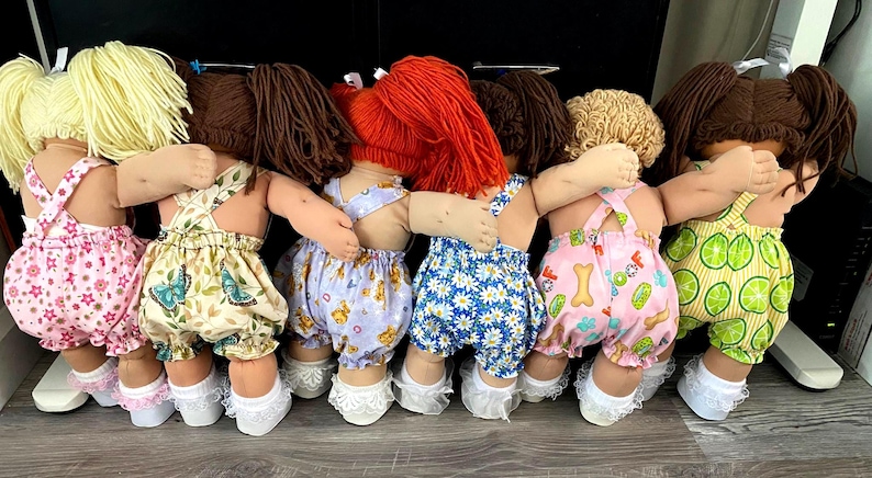 Puede incluir: Seis mu&ntilde;ecas de pie en fila con los brazos alrededor de las otras. Cada mu&ntilde;eca tiene pelo de lana amarillo, marr&oacute;n o rojo y viste petos de tela estampada con calcetines blancos con volantes. Los conjuntos tienen estampados de flores y frutas.