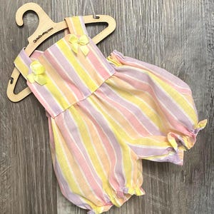 Puede incluir: Un pelele sin mangas con rayas verticales en tonos pastel de amarillo, rosa y lavanda. La prenda tiene un escote cuadrado, dos peque&ntilde;os lazos amarillos con cuentas centrales en los hombros y aberturas de pierna fruncidas en una percha de madera.