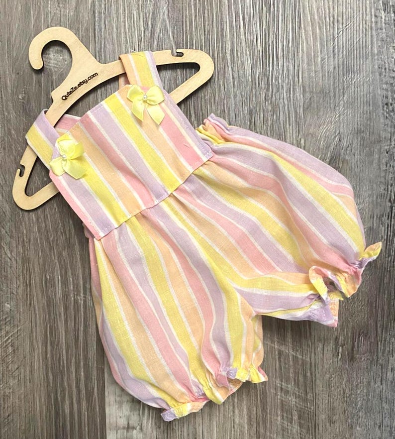Puede incluir: Un pelele sin mangas con rayas verticales en tonos pastel de amarillo, rosa y lavanda. La prenda tiene un escote cuadrado, dos peque&ntilde;os lazos amarillos con cuentas centrales en los hombros y aberturas de pierna fruncidas en una percha de madera.