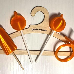 May include: A light-colored wooden hanger with the text QuteZe.etsy.com printed on it, featuring four orange plastic accessories: a cylindrical piece, two circular pieces on white sticks, and a ring-shaped piece with a small protruding tab.