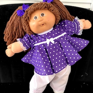 May include: A doll with brown yarn hair styled in two pigtails with purple bows. The doll wears a purple dress with white polka dots, a white ribbon bow at the waist, and white lace trim on the sleeves, paired with plain white long pants.