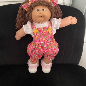 Puede incluir: Una mu&ntilde;eca con cabello de lana marr&oacute;n peinado en coletas con lazos florales a juego. La mu&ntilde;eca viste una blusa blanca de manga corta con cuello de encaje bajo un peto floral rosa con detalles de lazos amarillos, calcetines blancos con encaje y zapatos blancos.