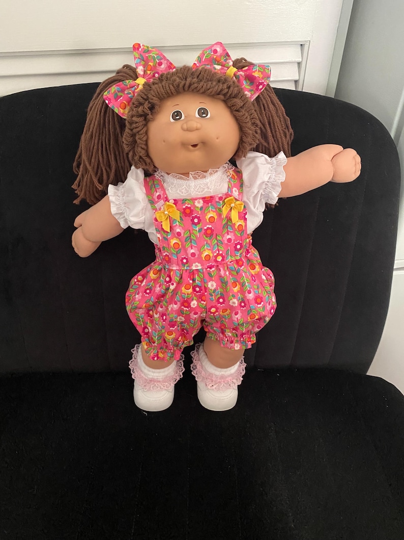 Puede incluir: Una mu&ntilde;eca con cabello de lana marr&oacute;n peinado en coletas con lazos florales a juego. La mu&ntilde;eca viste una blusa blanca de manga corta con cuello de encaje bajo un peto floral rosa con detalles de lazos amarillos, calcetines blancos con encaje y zapatos blancos.