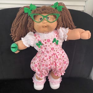 Puede incluir: Una mu&ntilde;eca con cabello de lana marr&oacute;n peinado en coletas con lazos verdes, gafas verdes de ojo de gato, una blusa blanca de manga corta con encaje y un peto rosa con estampado floral y peque&ntilde;os lazos verdes. La mu&ntilde;eca lleva zapatos blancos.