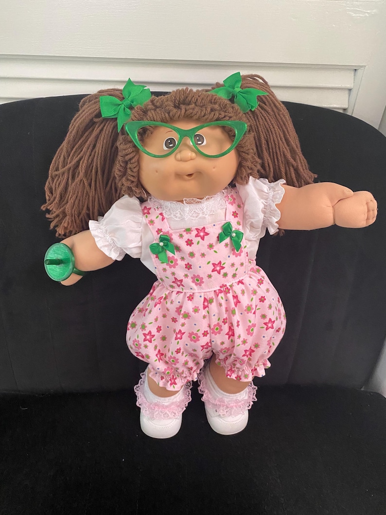 Puede incluir: Una mu&ntilde;eca con cabello de lana marr&oacute;n peinado en coletas con lazos verdes, gafas verdes de ojo de gato, una blusa blanca de manga corta con encaje y un peto rosa con estampado floral y peque&ntilde;os lazos verdes. La mu&ntilde;eca lleva zapatos blancos.