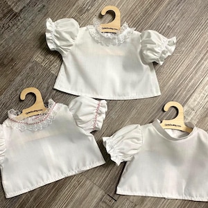 Puede incluir: Tres camisas blancas de manga corta abullonada, cada una expuesta en una peque&ntilde;a percha de madera. Una camisa tiene un cuello de encaje blanco, otra un cuello de encaje blanco con costuras rojas y la tercera un cuello alto sencillo.