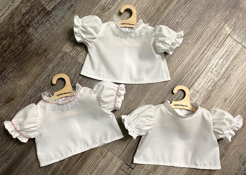 Puede incluir: Tres camisas blancas de manga corta abullonada, cada una expuesta en una peque&ntilde;a percha de madera. Una camisa tiene un cuello de encaje blanco, otra un cuello de encaje blanco con costuras rojas y la tercera un cuello alto sencillo.