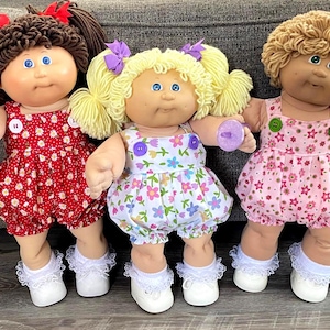 May include: Three Cabbage Patch Kids dolls. The dolls have different hair colors and styles, and wear dresses with floral patterns. Each doll has white shoes with lace trim and a pacifier. The doll on the left has brown hair and a red dress.