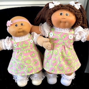 May include: Two Cabbage Patch Kids dolls. One doll is bald with a pink headband and a pink flower. The other doll has brown yarn pigtails with white bows. Both dolls wear white dresses with a pink and green frog print.