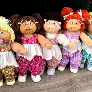 May include: Five Cabbage Patch Kids dolls standing in a row. Each doll has yarn hair, round plastic glasses, and wears a white-topped outfit with patterned pants. The first doll on the left holds a purple lollipop. They have soft fabric bodies.