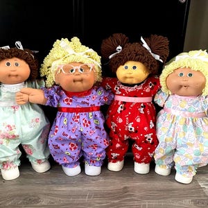 May include: Four Cabbage Patch Kids dolls wearing different outfits. The dolls are all wearing pajamas. The dolls are all smiling.