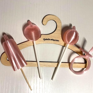 May include: A light wooden clothes hanger with the text QuteZe.etsy.com printed on it, featuring three pink, translucent, plastic-like items attached to it: a cylindrical object, two round lollipop-shaped pieces, and a ring-shaped pacifier toy.