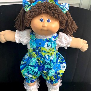 Puede incluir: Una mu&ntilde;eca con cabello de lana marr&oacute;n peinado en coletas y un lazo a juego con estampado floral azul y verde. La mu&ntilde;eca viste una blusa blanca de manga corta bajo un peto floral azul y verde, calcetines blancos con encaje y zapatos blancos.