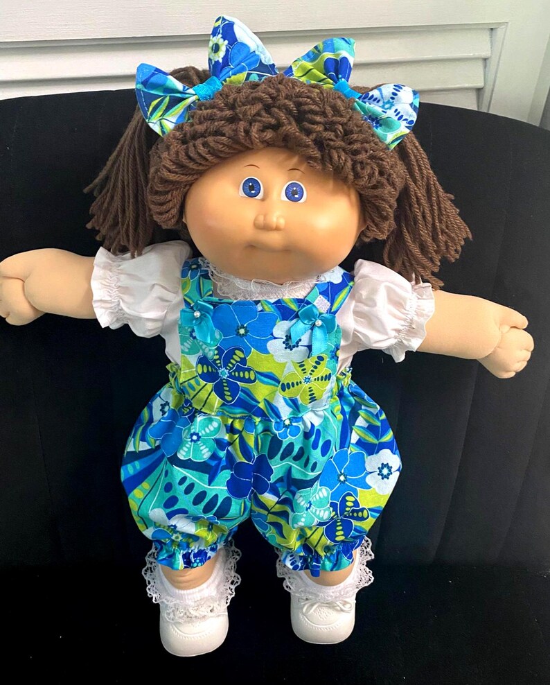 Puede incluir: Una mu&ntilde;eca con cabello de lana marr&oacute;n peinado en coletas y un lazo a juego con estampado floral azul y verde. La mu&ntilde;eca viste una blusa blanca de manga corta bajo un peto floral azul y verde, calcetines blancos con encaje y zapatos blancos.