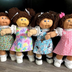 May include: Four vintage Cabbage Patch Kids dolls. Each doll has brown yarn pigtails, a round face, and is wearing a dress. The dresses are in various patterns, including floral, butterfly, and anchor prints. The dolls are wearing white shoes.