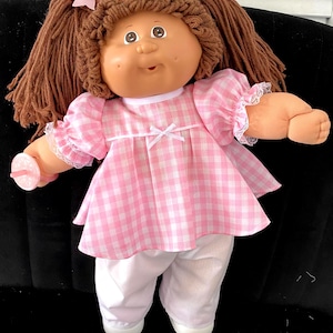 May include: A vintage doll with brown yarn hair styled in pigtails, adorned with pink bows. The doll wears a pink and white checkered dress with lace trim and white pants. It holds a pink pacifier.