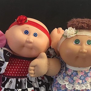 May include: Four dolls with round faces and soft bodies. The first wears a pink dress and purple pacifier. The second has a red headband and black and white dress. The third has brown hair and a floral dress. The fourth wears a yellow headband and white pacifier.