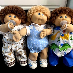 May include: Three Cabbage Patch Kids dolls, each with a pacifier. One doll wears a blue polka-dot romper, another wears a dinosaur-print shirt and blue pants, and the third wears dog-print pajamas. All have blue eyes and curly hair.