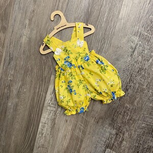 Puede incluir: Un pelele amarillo brillante con un estampado floral azul y blanco, aberturas el&aacute;sticas en las piernas y dos peque&ntilde;os lazos blancos en los tirantes. La prenda se muestra en una percha de madera clara con el texto QuteZu.etsy.com.