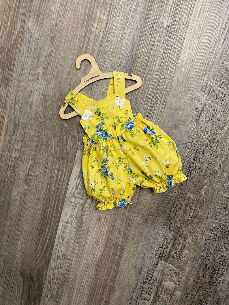 Puede incluir: Un pelele amarillo brillante con un estampado floral azul y blanco, aberturas el&aacute;sticas en las piernas y dos peque&ntilde;os lazos blancos en los tirantes. La prenda se muestra en una percha de madera clara con el texto QuteZu.etsy.com.
