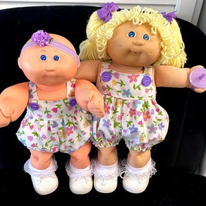 May include: Two vintage Cabbage Patch Kids dolls. One doll has a bald head with a purple headband and the other has blonde curly hair. Both dolls wear floral print rompers with purple buttons, white shoes, and lace-trimmed socks. One doll holds a purple accessory.