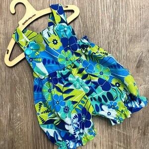 Puede incluir: Un pelele sin mangas con un vibrante estampado floral en tonos azul, verde lima y blanco. La prenda tiene aberturas el&aacute;sticas en las piernas, un escote cuadrado y dos peque&ntilde;os lazos de sat&eacute;n azul con cuentas blancas en los hombros.