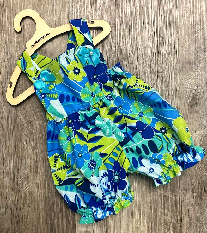 Puede incluir: Un pelele sin mangas con un vibrante estampado floral en tonos azul, verde lima y blanco. La prenda tiene aberturas el&aacute;sticas en las piernas, un escote cuadrado y dos peque&ntilde;os lazos de sat&eacute;n azul con cuentas blancas en los hombros.